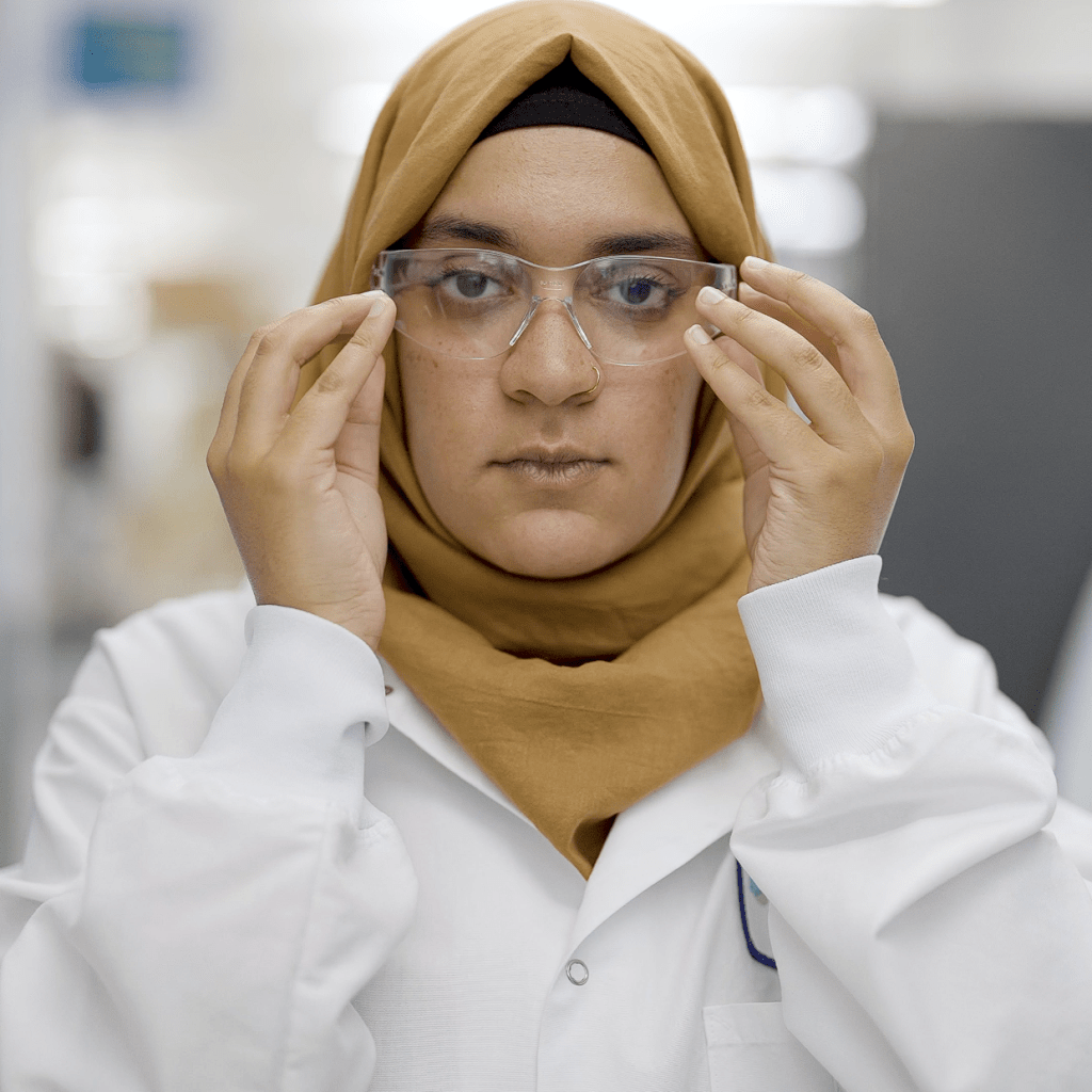 A female scientist with a lab coat on puts on safety glasses. She is wearing a brown hijab.